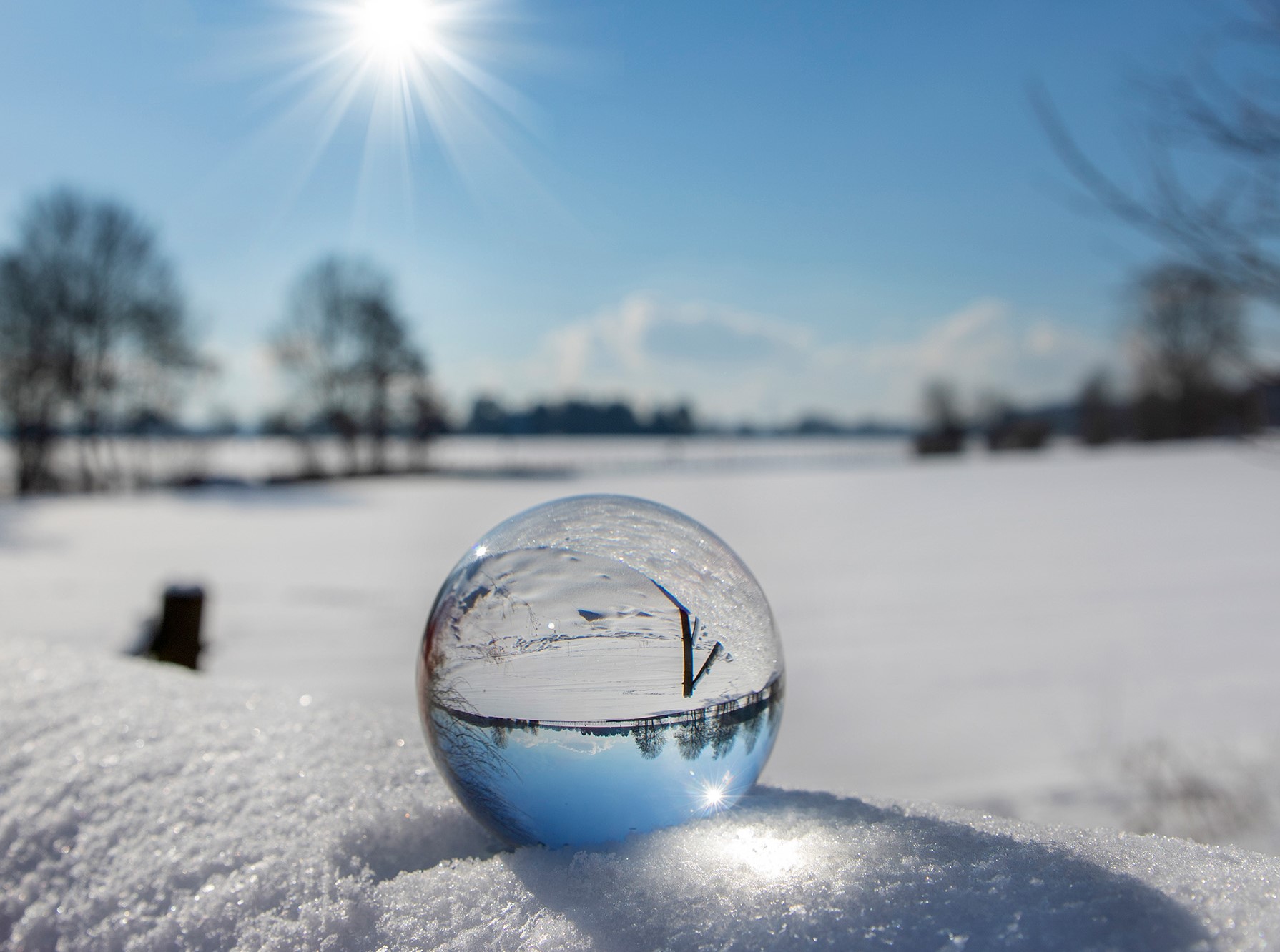 Glaskugel liegt in schneebedeckter Landschaft. Der Himmel und die Sonnenstrahlen spiegeln sich in der Glaskugel.